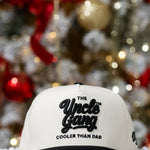 White cap with 'The Uncle Gang Cooler Than Dad' text in front of a decorated Christmas tree.