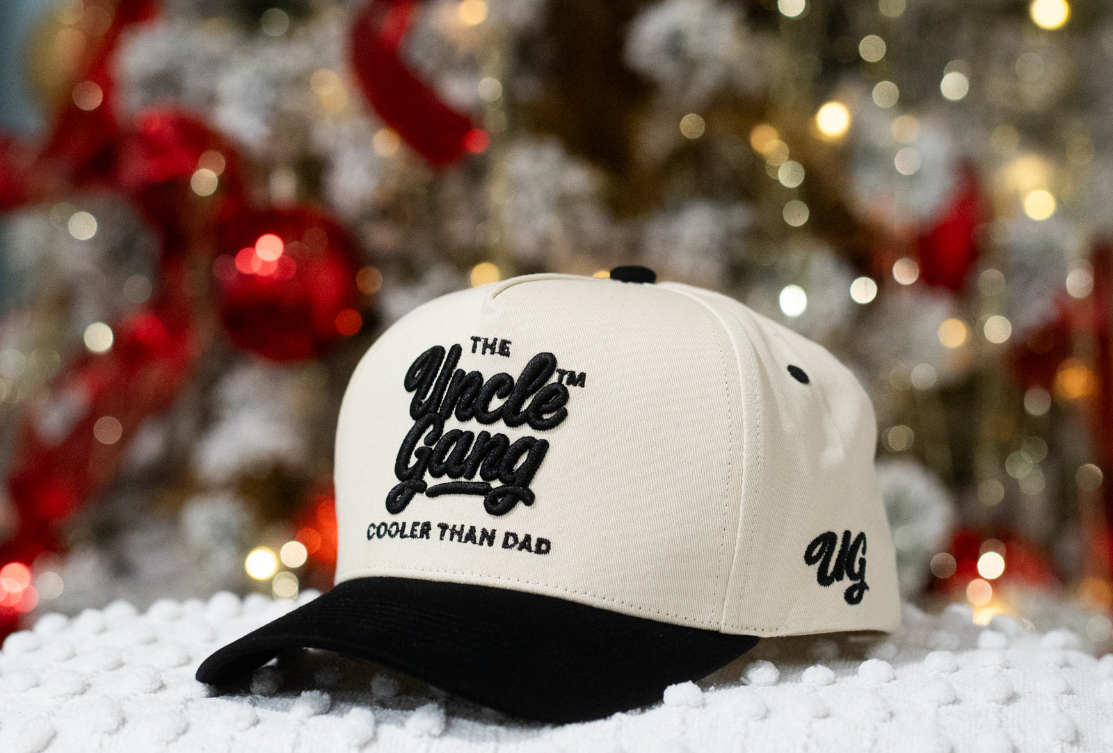 Cap with 'The Uncle Gang Cooler Than Dad' text on a snowy surface with blurred Christmas tree in the background