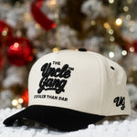 Cap with 'The Uncle Gang Cooler Than Dad' text on a snowy surface with blurred Christmas tree in the background