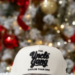 White cap with 'The Uncle Gang Cooler Than Dad' text in front of a decorated Christmas tree.